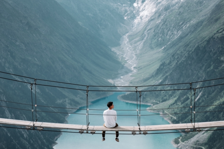 Man zit op hangbrug tussen twee bergen en kijkt naar een meer. Foto: Alex Azabache via Unsplash